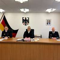 Eine Richterin sitzt zwischen zwei Soldaten an einem Tisch vor Unterlagen und Büchern