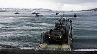 Ein militärisches Fahrzeug fährt von einem Boot runter, welches eine Klappe aufgeklappt hat.