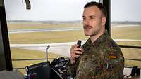 Ein Soldat steht mit einem Mikrofon in der Hand in einem Tower. Im Hintergrund ein Flugplatz.