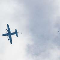 Silhouette einer C-130J von unten unter Wolken mit Fallschirmen