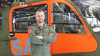 Ein Soldat in Fliegeruniform steht vor einem Hubschrauber mit orangefarbener Lackierung.