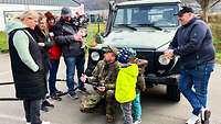 Ein Soldat hockt vor stehenden Zivilisten, hält einen kleinen Monitor in den Händen und spricht
