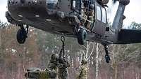 Unter einem schwebenden Hubschrauber stehen Soldaten und halten eine Schlaufe hoch