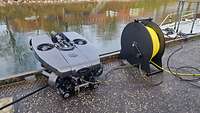 A black device at the water’s edge with a yellow tether on a reel to its right.