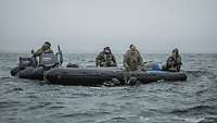 Mehrere Soldaten sitzen in einem Schlauchboot was im Wasser schwimmt, Taucher halten sich daran fest