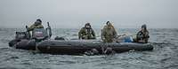 Mehrere Soldaten sitzen in einem Schlauchboot was im Wasser schwimmt, Taucher halten sich daran fest