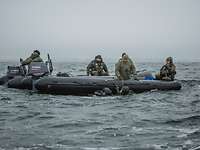 Mehrere Soldaten sitzen in einem Schlauchboot was im Wasser schwimmt, Taucher halten sich daran fest