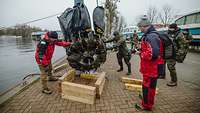 Ein Flugzeugmotor wird mit einem Kran sowie Führungsseilen von mehreren Personen aus dem Wasser geborgen