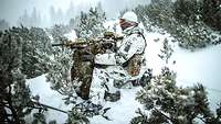 Ein Soldat mit Maschinengewehr hockt zwischen kleinen Bäumen im verschneiten Gebirge.