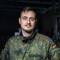 Ein Mann in Uniform steht vor einem dunklen Hintergrund. 