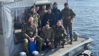 Mehrere Soldaten stehen vor einem grauen Zelt. Um sie herum ist ein Gewässer zu erkennen.