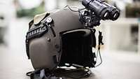 A helmet is lying on the ground. A small pair of binoculars is attached to the top of the helmet.