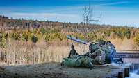 Drei Soldaten hocken auf einer Wiese und schießen eine Panzerfaust in die Luft