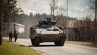 Ein Schützenpanzer vom Typ Marder fährt eine Straße entlang, zwei Soldaten stehen am Seitenrand