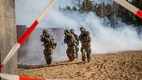 Vier Soldaten bewegen sich mit Waffe im Anschlag über eine sandige Fläche auf ein Gebäude zu, hinter ihnen eine Nebelwolke