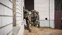 Drei Soldaten stehen nah vor einem Gebäude. Vor ihnen klettert ein vierter Soldat durch eine Fensteröffnung in das Gebäude