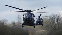 Zwei Hubschrauber schweben hintereinander in der Luft. Hinter ihnen alte Lagerhallen.