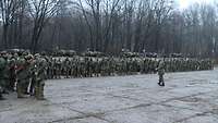 Soldaten treten an. Hinter ihnen stehen mehrere Schützenpanzer und davor ein Soldat.