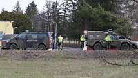 Zwei Fahrzeuge verschiedener Nationen stehen am Straßenrand. Davor stehen mehrere Soldaten.