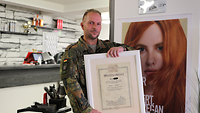 Ein Soldat hält seinen Meisterbrief in den Händen. Er steht in einem Beautysalon.