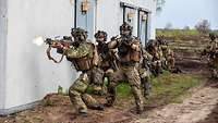 Mehrere Soldaten stehen oder hocken hintereinander an einer Hauswand, der Soldat vorn schießt. 