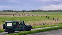 Mehrere Soldaten laufen nebeneinander über eine Wiese. Im Vordergrund steht ein Militärfahrzeug.