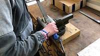 In einem Schraubstock ist ein Stück Metall eingeklemmt. Zwei Hände halten darüber eine Metallfeile.