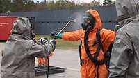 Mensch im grauen Schutzanzug spritzt eine Person im orangenen Overall mit Atemschutzmaske ab