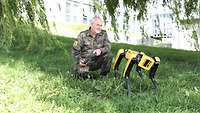 Ein Mann in Uniform kniet neben einem gelb-schwarzen Roboter auf vier Beinen auf einer Wiese