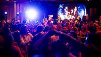 Dicht gedrängt tanzen Menschen bei schummrigem Licht, vorn spielt eine Band und zwei singen.
