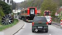 Schwerlasttransport von hinten: Begleitfahrzeuge, die eine schmale Dorfstraße in einem Tal passieren