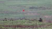 Zwei Soldaten im Gelände auf und vor ihrem Schützenpanzer, von dem nur das Geschützrohr zu sehen ist