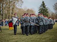 Soldaten marschieren in Formation über eine Wiese, vorn eine Soldatin mit weißer Dreiecksfahne