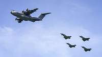 Ein graues, viermotoriges Transportflugzeug und vier Kampfjets vor einem leicht bewölkten Himmel.