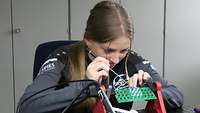 A young woman sitting on a workbench pointing at a PCB with a screwdriver.