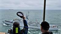 Ein graues Kriegsschiff in See; im Vordergrund winkt ein Mann mit einer weißen Schirmmütze.