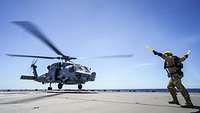 Ein Hubschrauber landet auf dem Flugdeck eines Schiffs, davor ein Soldaten mit gelben Stäben in den Händen