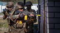 Drei Soldaten mit ihren Waffen im Anschlag laufen hintereinander an einer Mauer entlang.