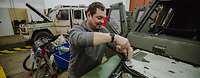 Eine Person schraubt an einem Fahrzeug in einer Halle, hinter ihm steht ein weiteres Fahrzeug