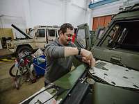 Eine Person schraubt an einem Fahrzeug in einer Halle, hinter ihm steht ein weiteres Fahrzeug