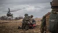 Mehrere Soldaten warten mit einem Verwundeten auf der Trage, im Hintergrund landet ein Hubschrauber