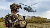 Ein Soldat steht in voller Montur mit einem Gewehr auf einem Feld, während ein Hubschrauber landet