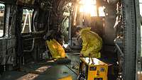 Two people dressed in yellow perform maintenance work in a helicopter.