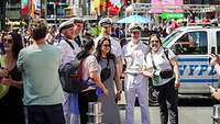 Several women pose for a cell phone photo next to sailors in white uniforms.