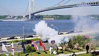 There are artillery pieces on a river bank with smoke rising above them.