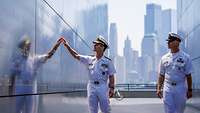 An admiral in a white uniform touches a polished stone wall with writing on it.