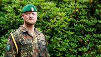 Ein Soldat steht vor einem Kompaniegebäude, rechts neben ihm steht ein Kompaniewappen aus Holz.