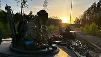 Zwei Soldaten schauen aus den Luken eines Transportpanzers auf einer Lichtung auf den Sonnenuntergang