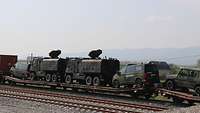 Ein Zugwagon auf dem mehrere Gefechtsfahrzeuge stehen, im Hintergrund sind Berge zu sehen