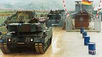 Mehrere Kampfpanzer im Konvoi fahren aus einem Feldlager, im Hintergrund sind Berge zu sehen